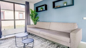 A room with a beige sofa, two round tables, a large window with curtains, and two wall-mounted shelves with plants.