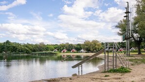 Järvenrantainen alue, jossa on metallinen liukumäki, hiekkaranta ja taustalla näkyviä taloja.