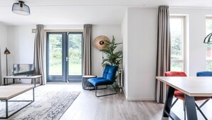 Uma sala de estar moderna com televisão, uma cadeira azul, uma mesa de jantar e uma janela grande com cortinas.