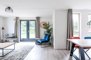 A modern living room with a television, a blue chair, a dining table, and a large window with curtains.