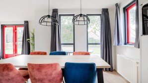 Uma sala de jantar com mesa e cadeiras, janelas amplas com cortinas e luminárias pendentes.