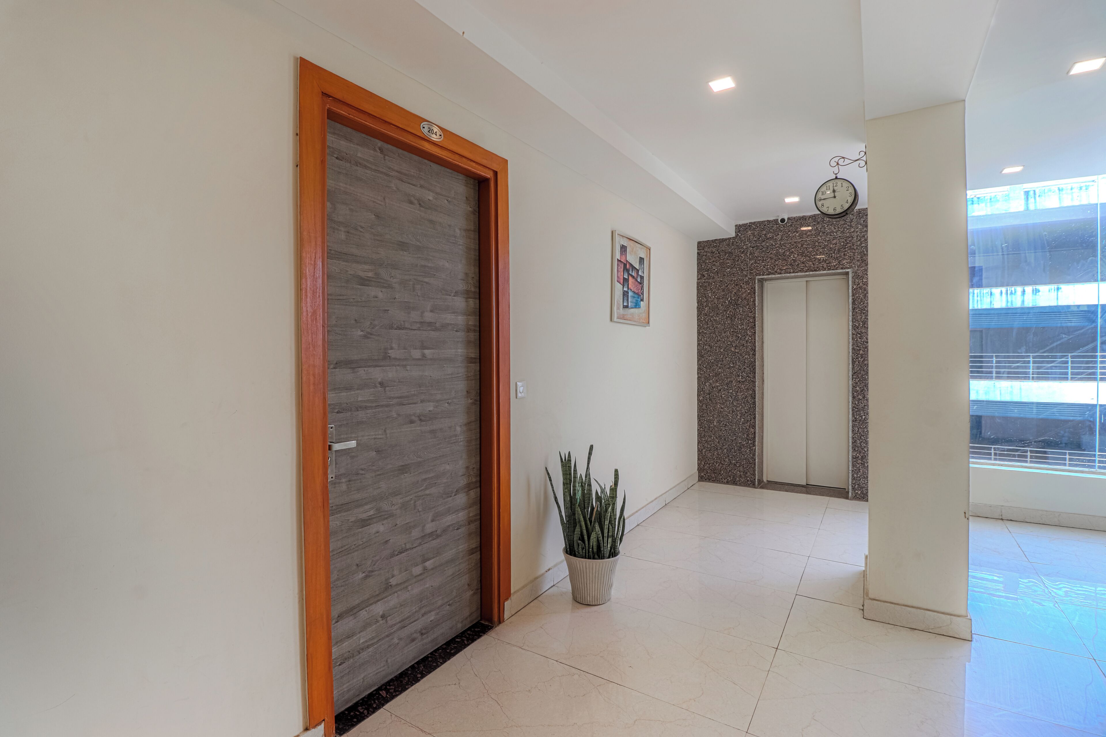 A hallway with a wooden door, a potted plant, a clock, and a door with a number plate.