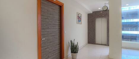 A hallway with a wooden door, a potted plant, a clock, and a door with a number plate.