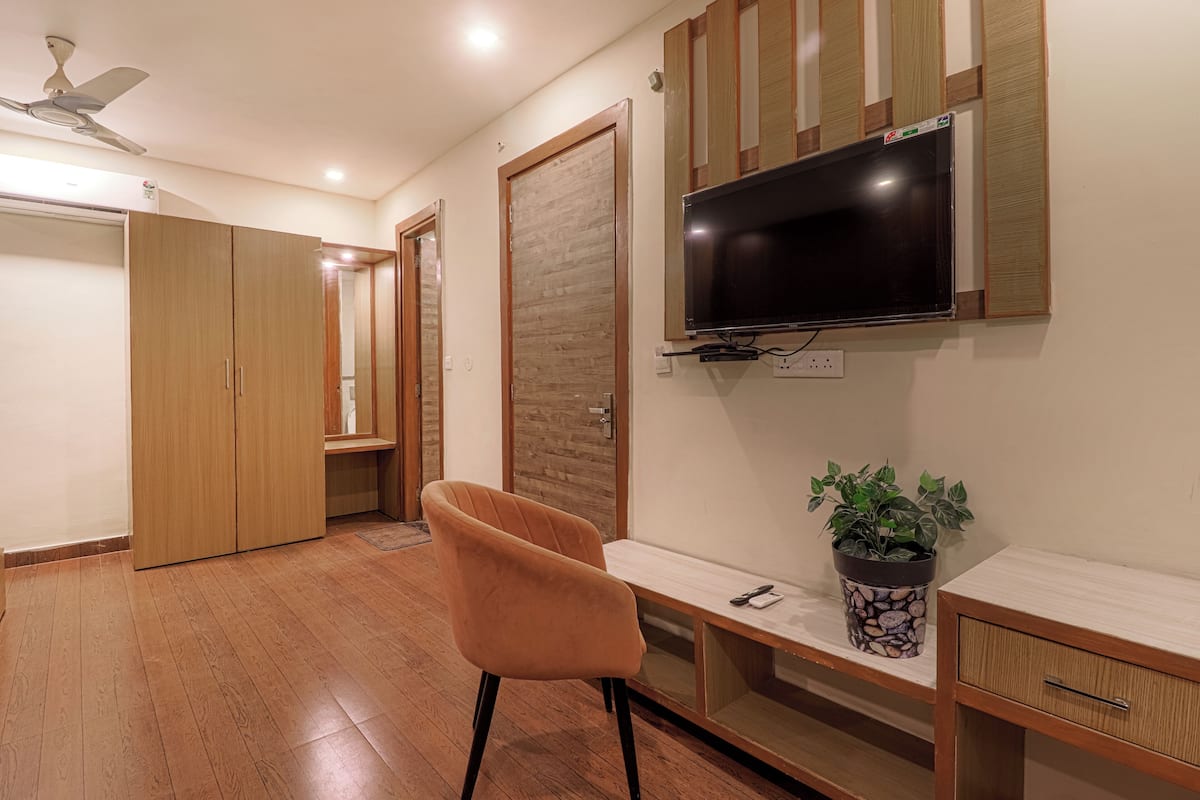 A room with a wooden floor, a ceiling fan, a TV mounted on the wall, a desk with a plant, and a chair.