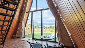 A room with a wooden ladder, two chairs, a small table with an open book, and a large window overlooking a green landscape and mountains. - Royal VIP Glamping Kintamani (Kintamani)