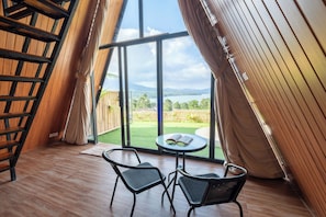 A room with a wooden ladder, two chairs, a small table with an open book, and a large window overlooking a green landscape and mountains.