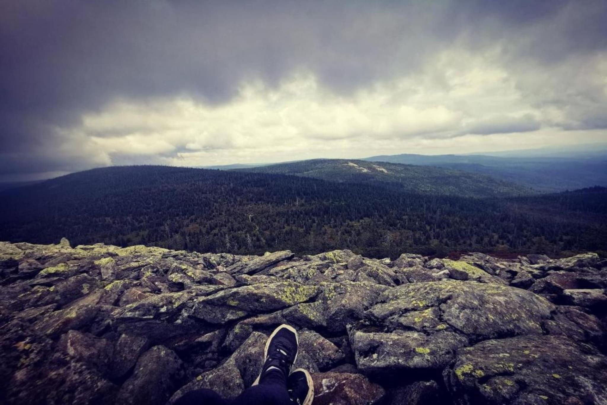 A rocky landscape with a person's feet on a rock, overlooking a forested valley under a cloudy sky.