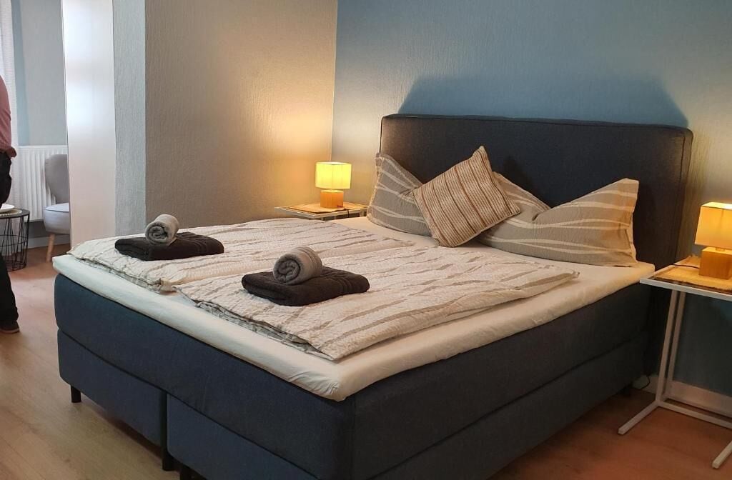 Una cama bien hecha con almohadas y una toalla doblada, una mesita de noche con una lámpara y una silla al fondo.