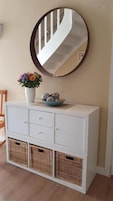 A white cabinet with wicker baskets, a vase with flowers, and a mirror reflecting a staircase.