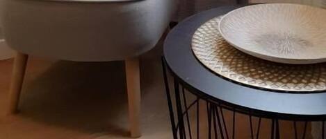 Una habitación con un sillón gris, una mesa redonda con tapa trenzada, un radiador y una ventana con cortinas.