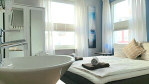 A hotel room with a large bathtub, a bed with pillows, and a window with curtains. - Seemoi (Norderney)
