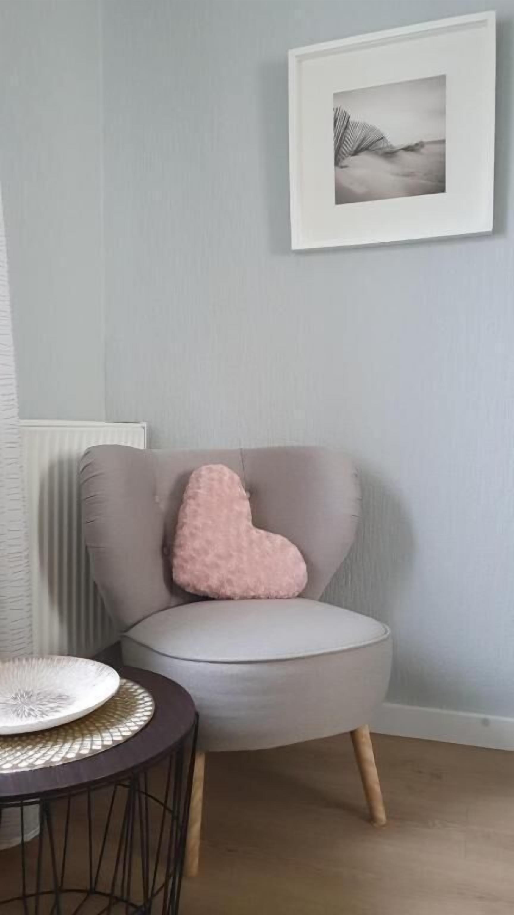A room with a round grey armchair, a small round table with a white plate, and a framed picture on the wall.