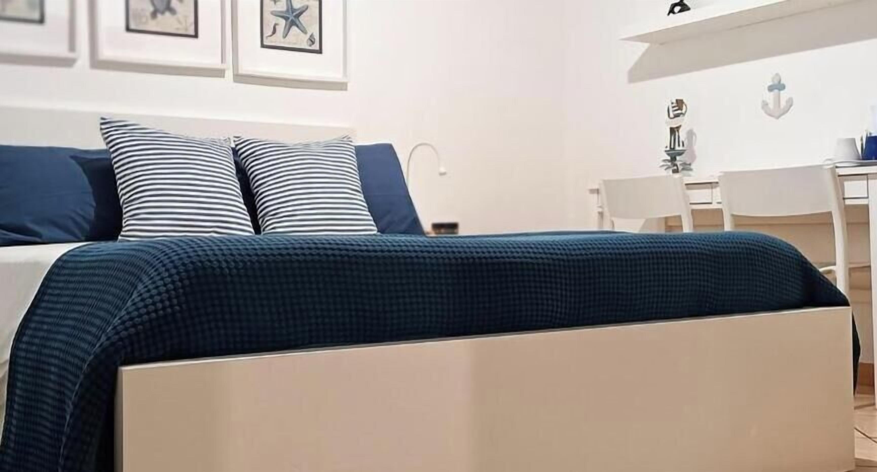 A neatly made bed with a striped pillow and a blue blanket, a white desk with a chair, and a wall with framed artwork.