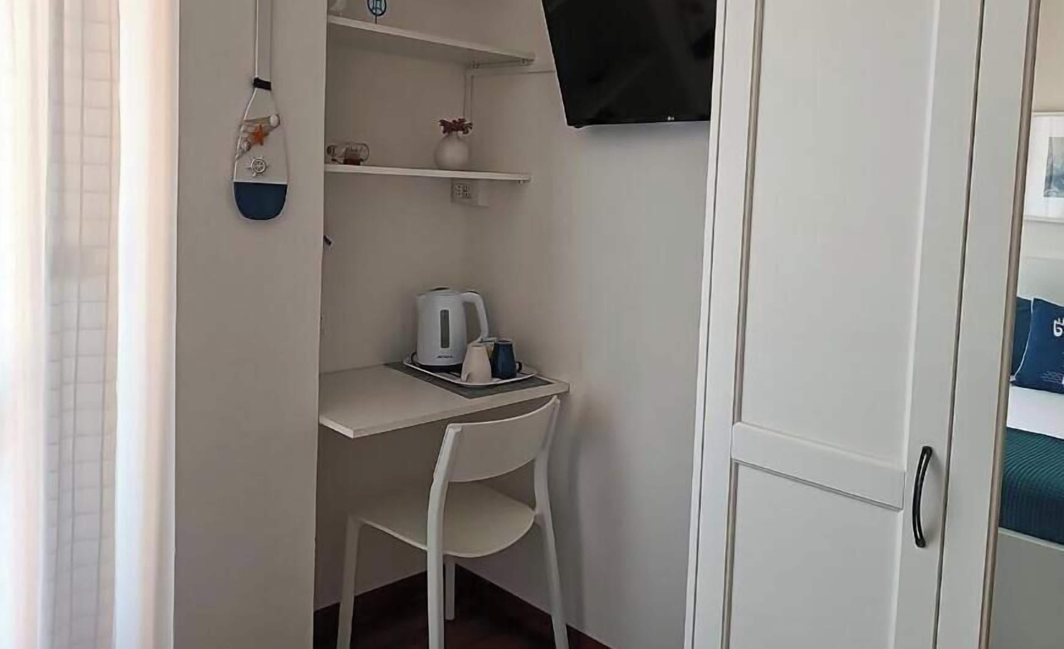 A small room with a white desk, a white chair, a kettle, and a TV mounted on the wall.