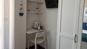 A small room with a white desk, a white chair, a kettle, and a TV mounted on the wall.