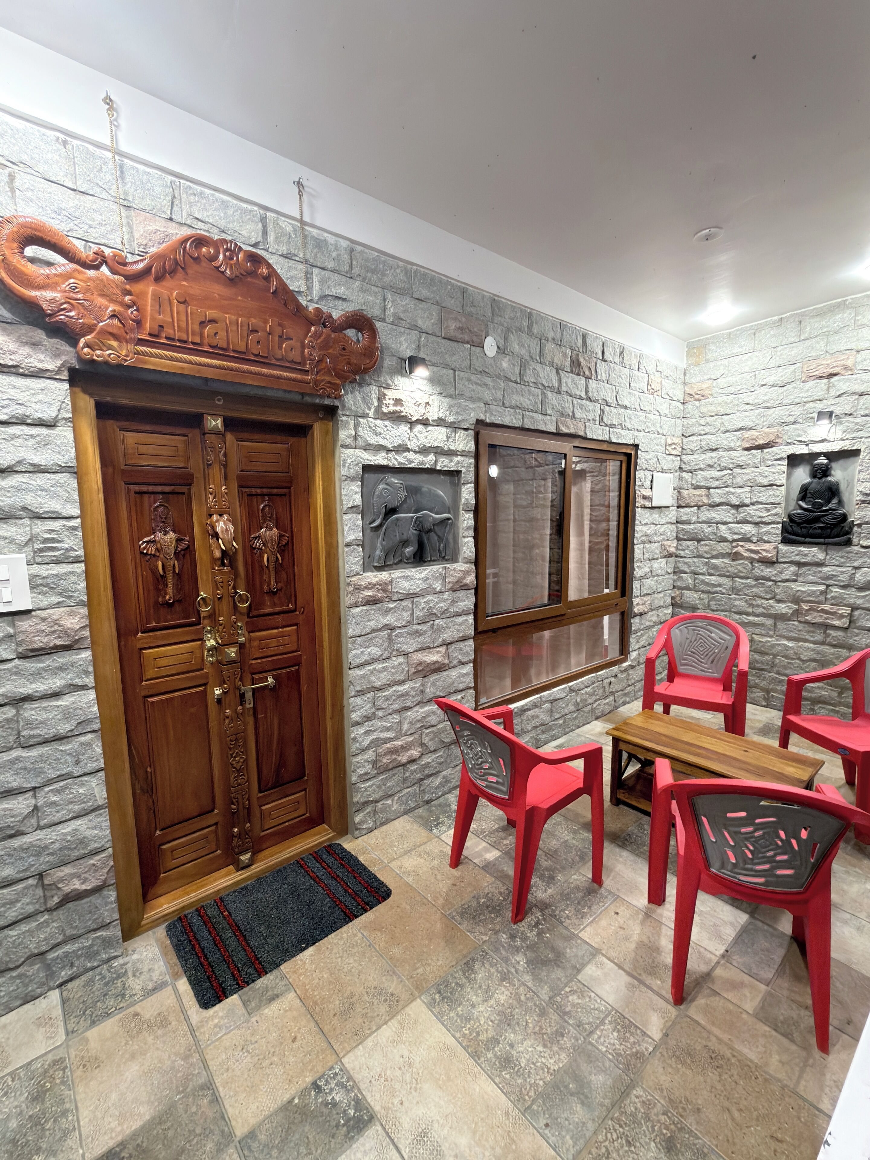 A room with a stone wall, a wooden door with intricate carvings, a small table with chairs, and a Buddha statue.
