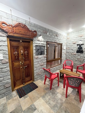 A room with a stone wall, a wooden door with intricate carvings, a small table with chairs, and a Buddha statue. - Deer Meadows  (Mudumalai)