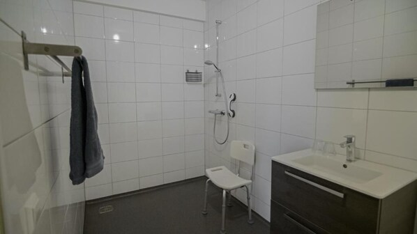 A bathroom with white tiled walls, a shower, a white chair, and a sink with a mirror above it.