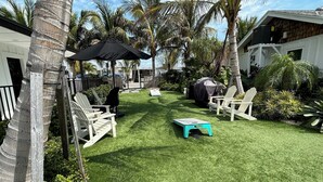 A well-maintained backyard with artificial grass, white Adirondack chairs, a small turquoise table, and a covered grill area. - Bridge Street Beach House | Wi-Fi | Center of All (Bradenton Beach)