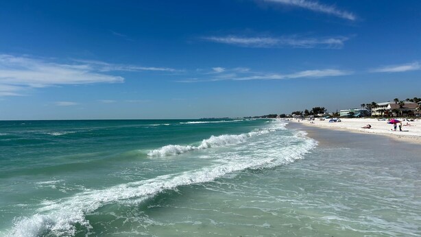 A beach with clear turquoise water and gentle waves, a sandy shore, and a few buildings in the background. - Bridge Street Beach House | Wi-Fi | Center of All (Bradenton Beach)