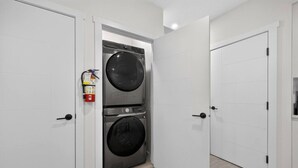 A laundry room with two stacked washing machines, white doors, and a fire extinguisher. - 2 Bed | Mins to Airport | Modern Suite (Calgary)