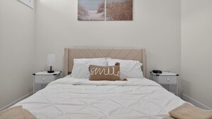 A bedroom with a bed, two bedside tables, a nightstand, a wall-mounted lamp, and two framed pictures. - 2 Bed | Mins to Airport | Modern Suite (Calgary)