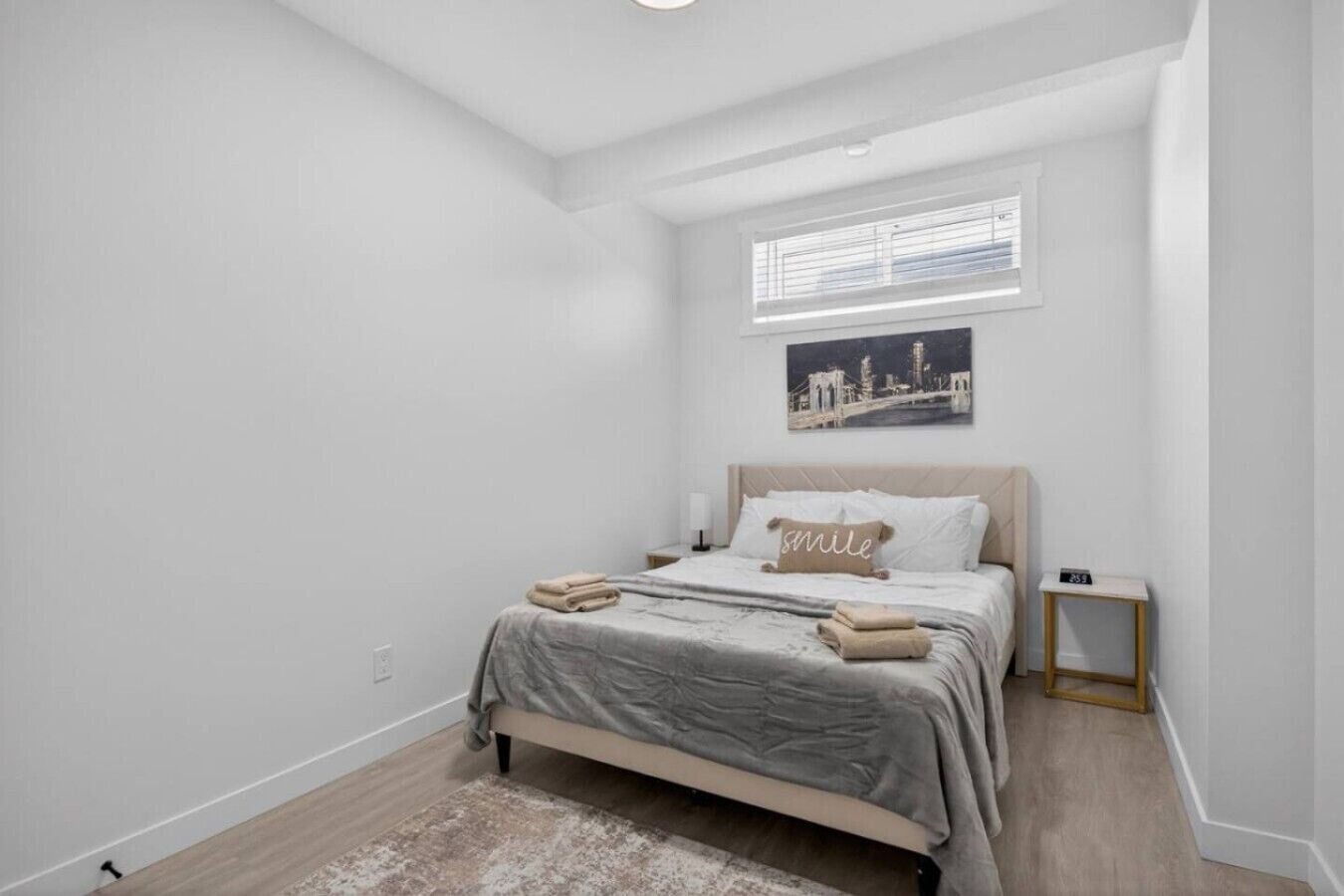 A bedroom with a bed, a nightstand, a small table, a window with blinds, and a cityscape painting on the wall.