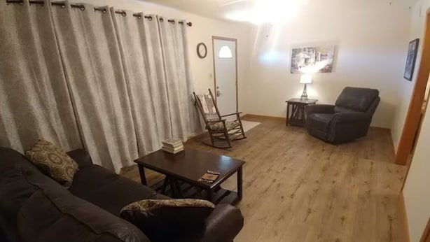 A living room with a dark brown sofa, a wooden coffee table, a wooden rocking chair, a black armchair, a small table with a lamp, and a painting on the wall.