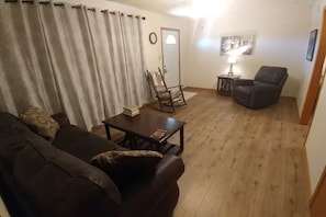 A living room with a dark brown sofa, a wooden coffee table, a wooden rocking chair, a black armchair, a small table with a lamp, and a painting on the wall. - 3-bedroom house in delightful Waynoka with cool AC breeze (Waynoka)