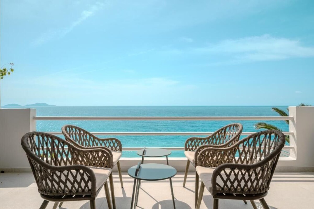 A terrace with wicker chairs and a small table, overlooking a clear blue sea.