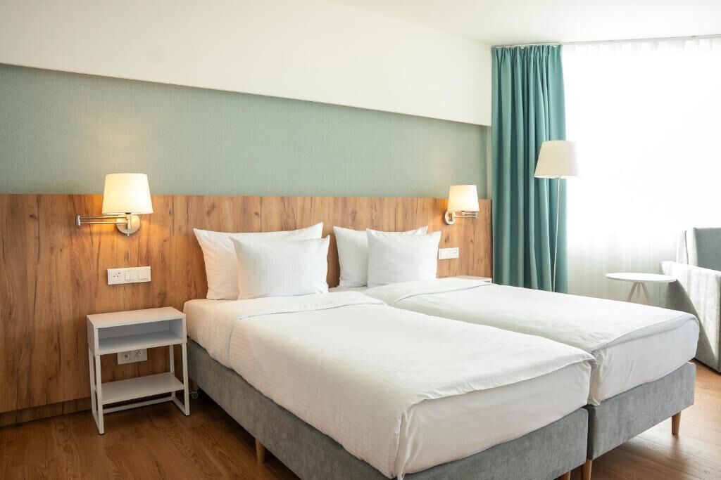 Ein Hotelzimmer mit zwei Betten, einem Holzkopfstück, Nachttischlampen, einem kleinen Tisch und Blick nach draußen.
