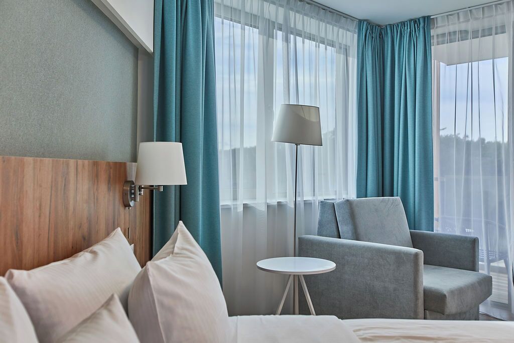 Habitación de hotel con cama, dos sillones, una mesita, lámpara y cortinas de encaje.