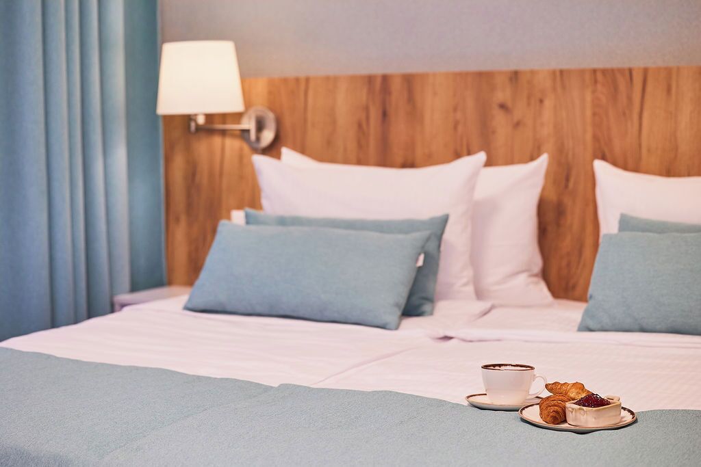 A hotel room with a wooden headboard, a bed with blue and white bedding, a bedside lamp, and a breakfast tray with croissants and a cup.