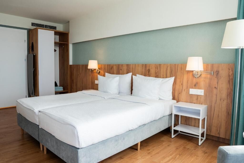 Una cama doble con sábanas blancas, un cabecero de madera, dos lámparas de noche, una mesita de noche pequeña y blanca, y un armario con espejo.