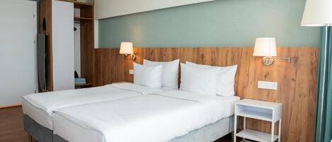 Una cama doble con sábanas blancas, un cabecero de madera, dos lámparas de noche, una mesita de noche pequeña y blanca, y un armario con espejo.