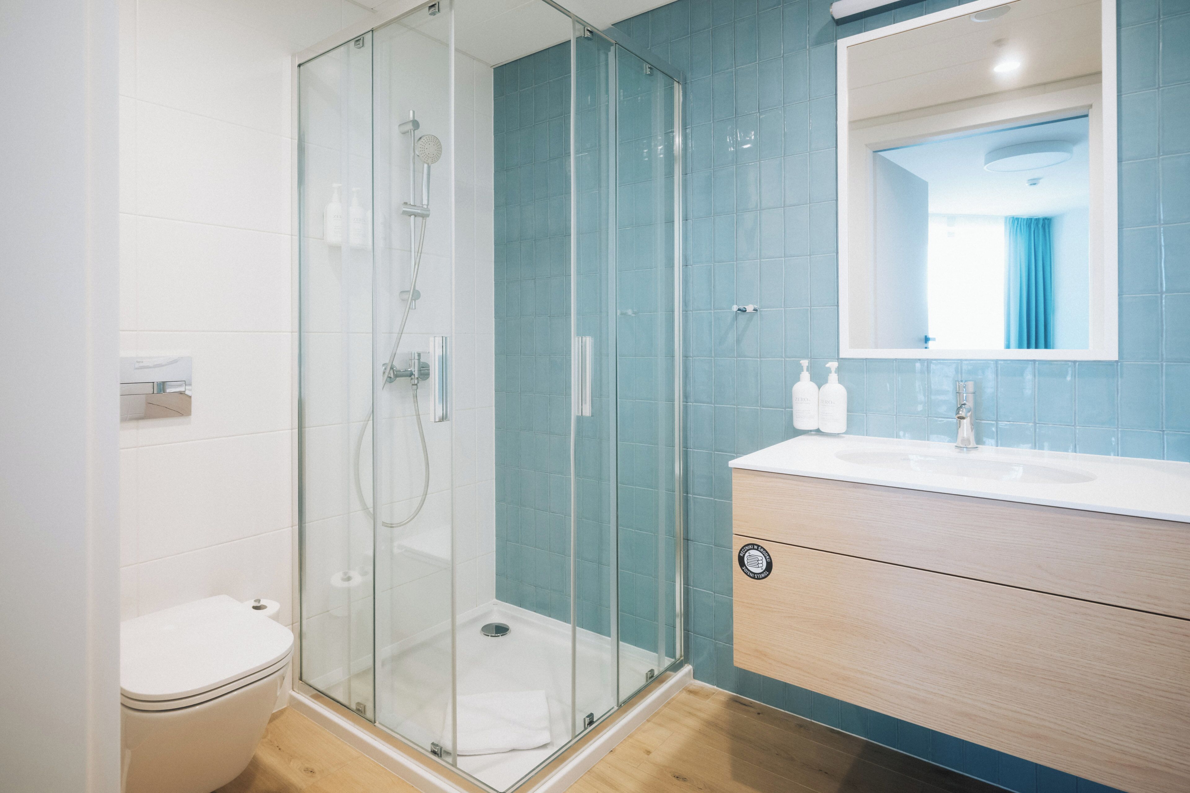 A modern bathroom with a glass shower enclosure, a white toilet, a wooden vanity with a mirror, and a blue-tiled wall.