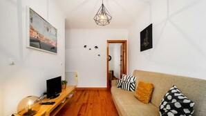 Una sala de estar con piso de madera, un sofá con cojines estampados, un televisor sobre un mueble de madera, una lámpara colgante y un cuadro en la pared.