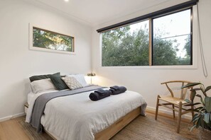 Ein Schlafzimmer mit Bett, Nachttisch, Lampe, Stuhl und einem Fenster mit Blick auf Bäume.