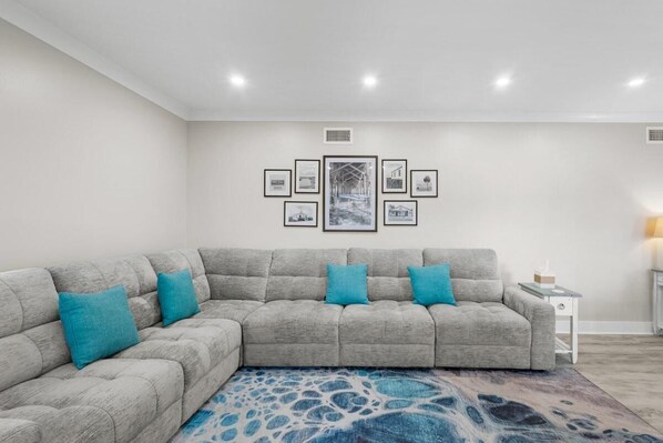 A modern living room with a grey sectional sofa, a patterned rug, and a wall-mounted art display. - Beach Time Oceanview 3 Bedroom (Pawleys Island)