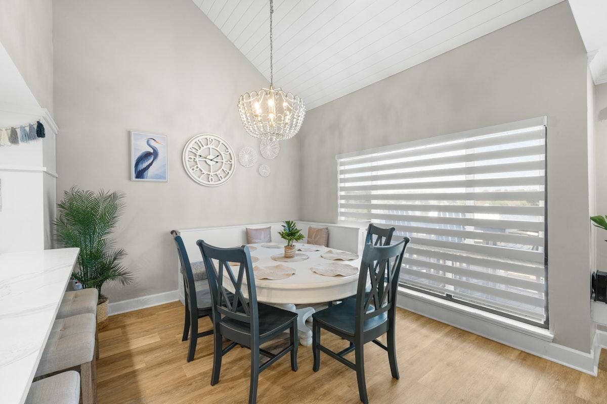 A dining area with a round table, four chairs, a chandelier, a large window with blinds, and wall decor.