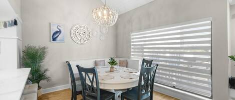 A dining area with a round table, four chairs, a chandelier, a large window with blinds, and wall decor.