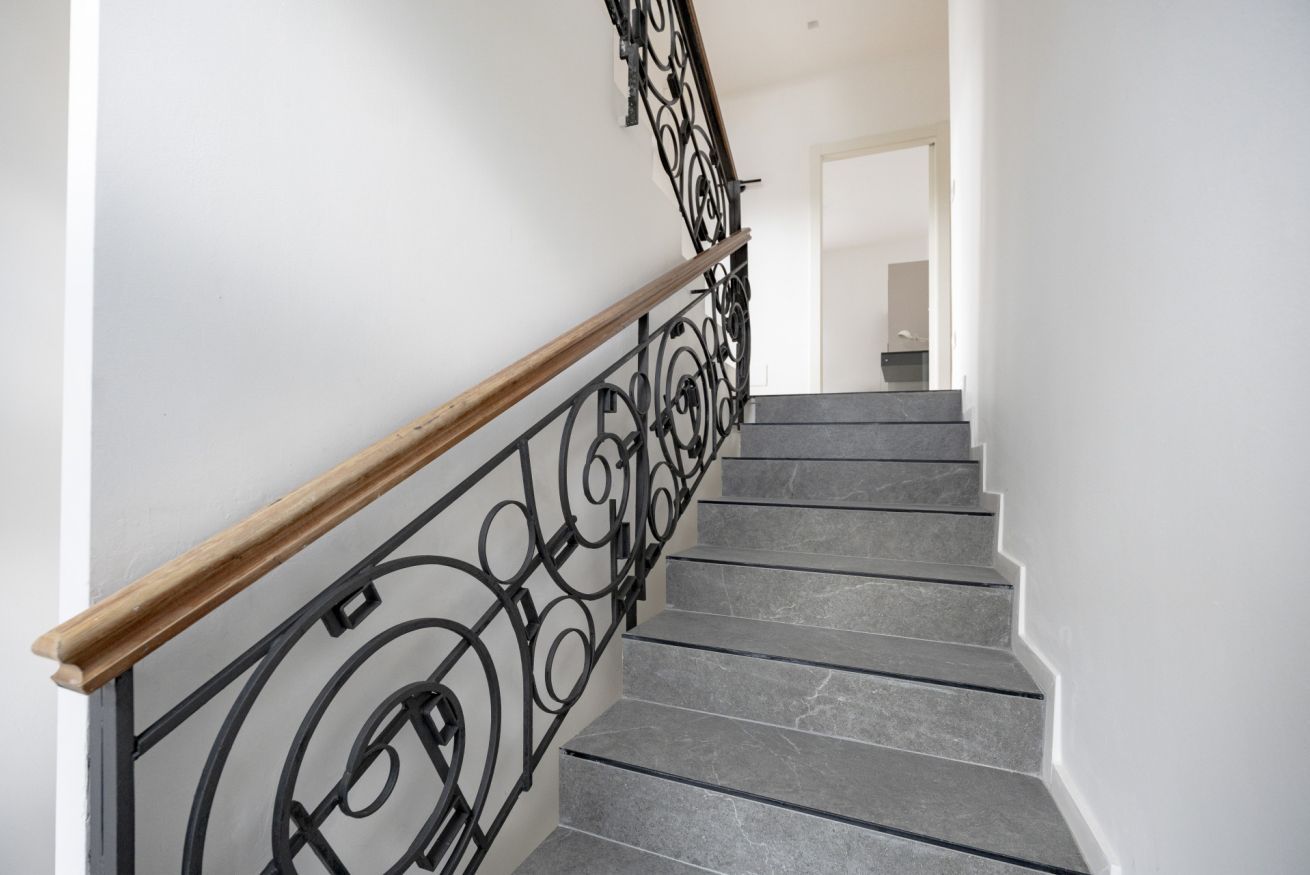 Uma escadaria com corrimão de madeira e balaústres de metal preto, que leva a um espaço interno.