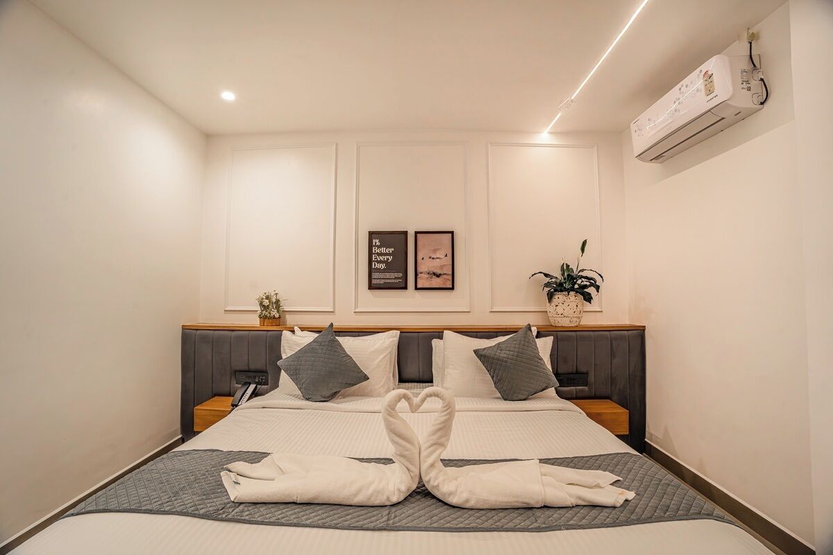 A hotel room with a bed, pillows, a bedside table, a wall-mounted air conditioner, and framed pictures.