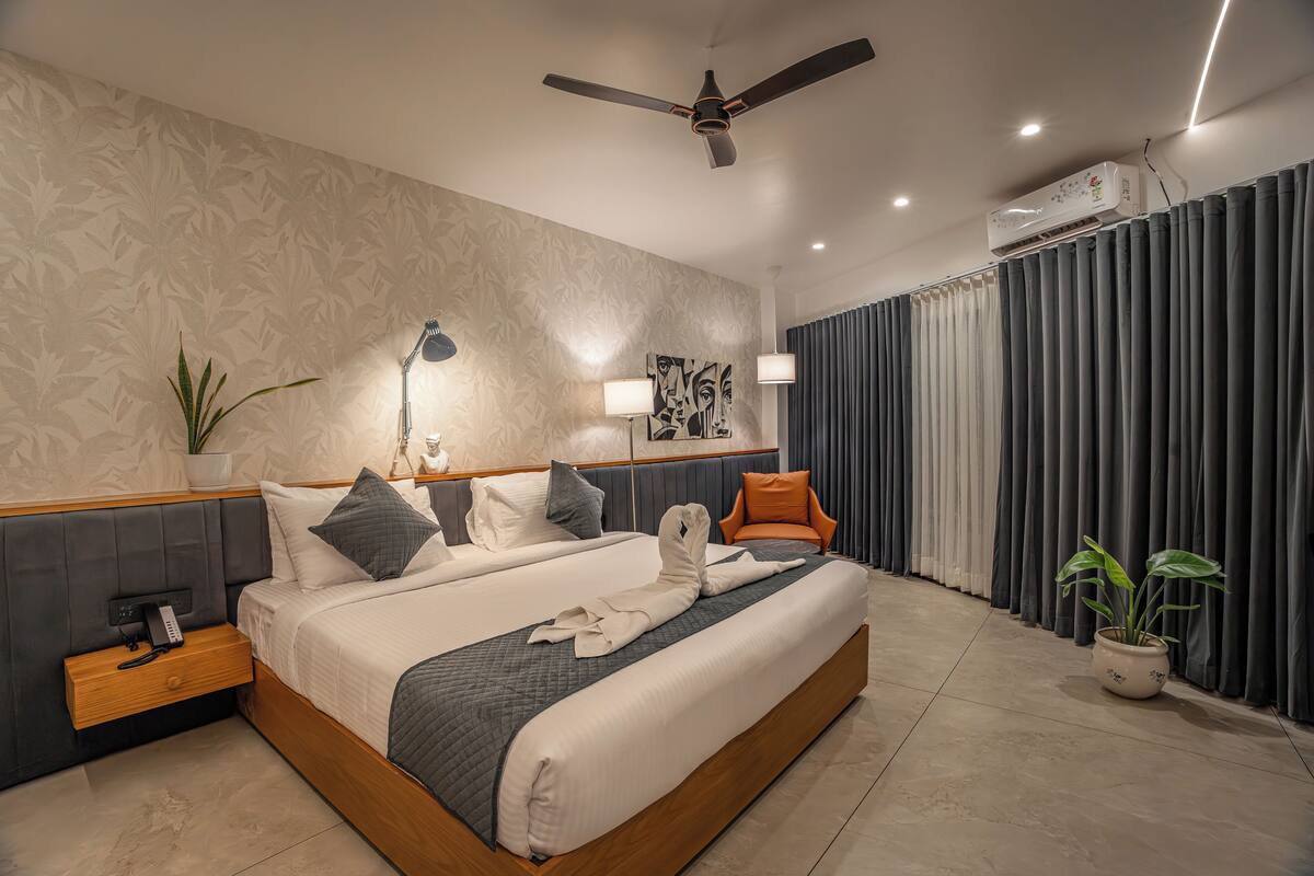 Quarto de hotel moderno com uma cama grande, mesas de cabeceira, ventilador de teto, aparelho de ar condicionado e um vaso de planta.