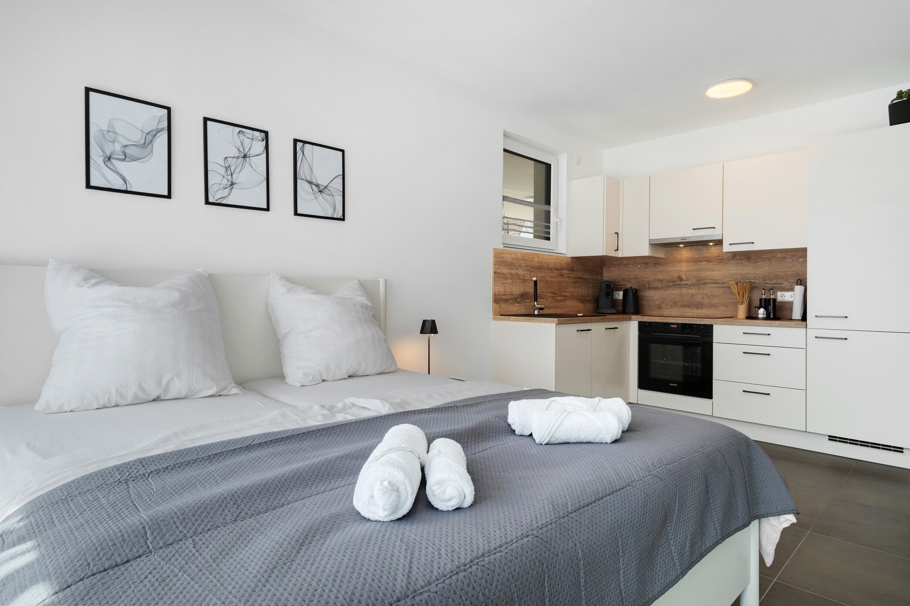 Una habitación de hotel moderna con una cama, una manta gris, almohadas blancas, una zona de cocina y tres obras de arte abstracto enmarcadas en la pared.