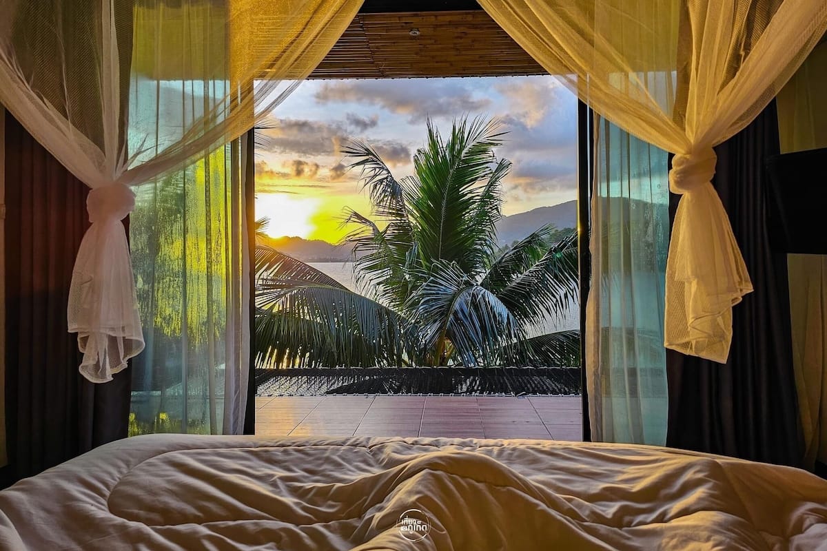 A bedroom with a bed, a view of palm trees and a sunset through sheer curtains.