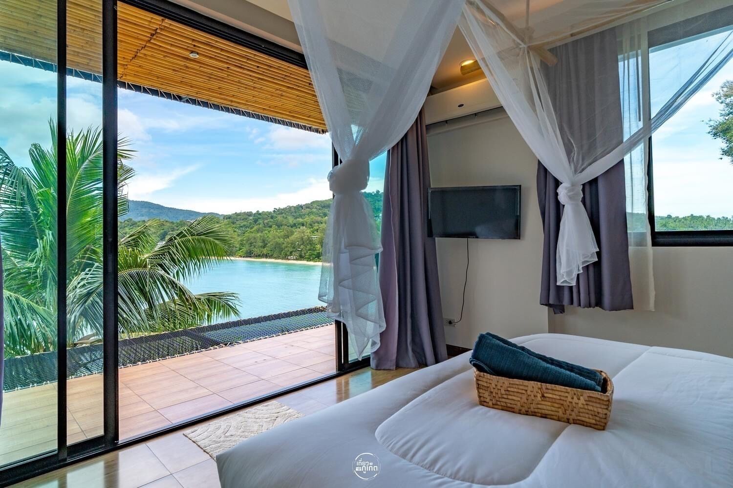 A bedroom with a large window, a bed, a TV, and a view of a beach and mountains.