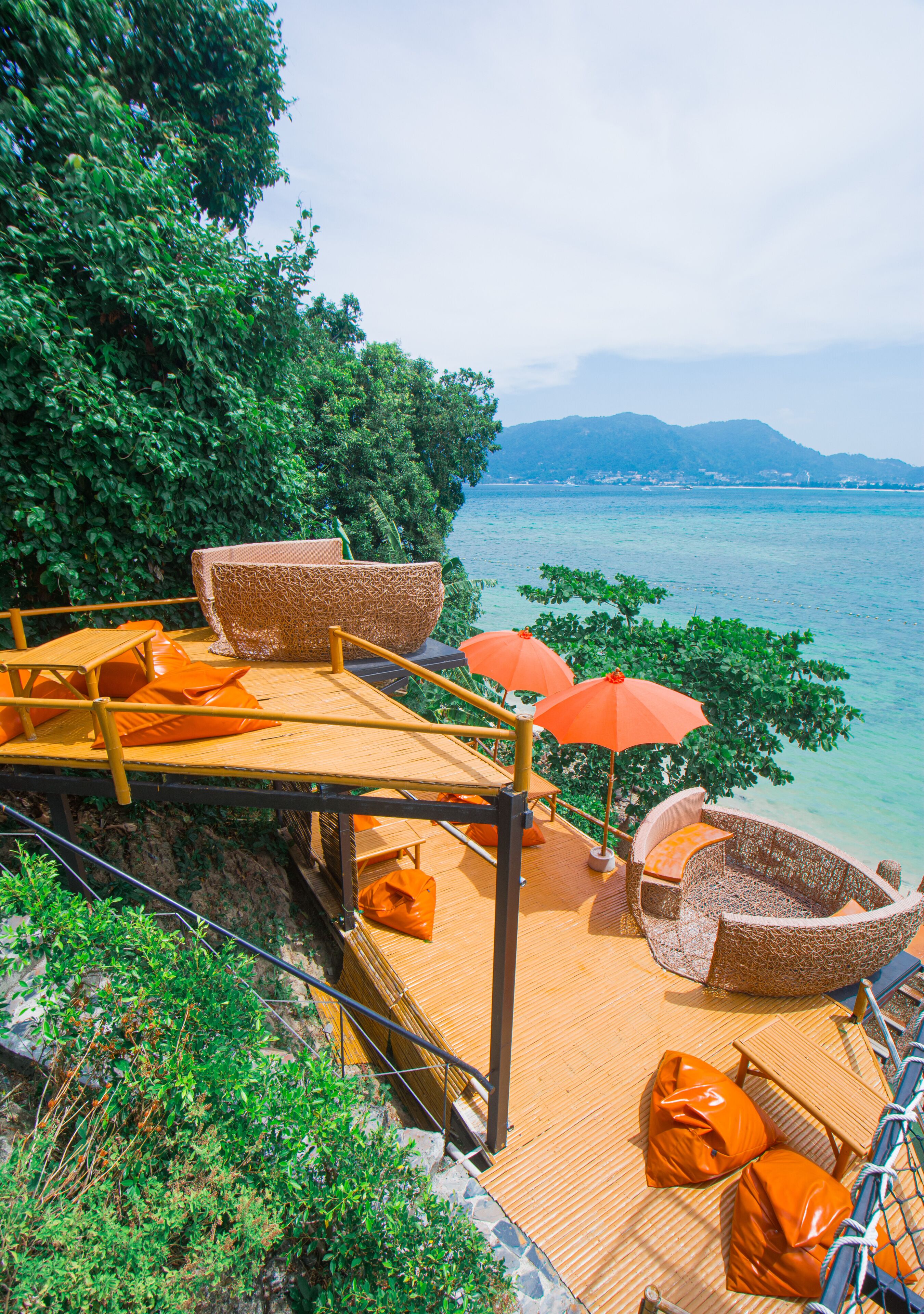 Eine Holzterrasse mit Korbmöbeln und orangefarbenen Kissen mit Blick auf ein Gewässer.