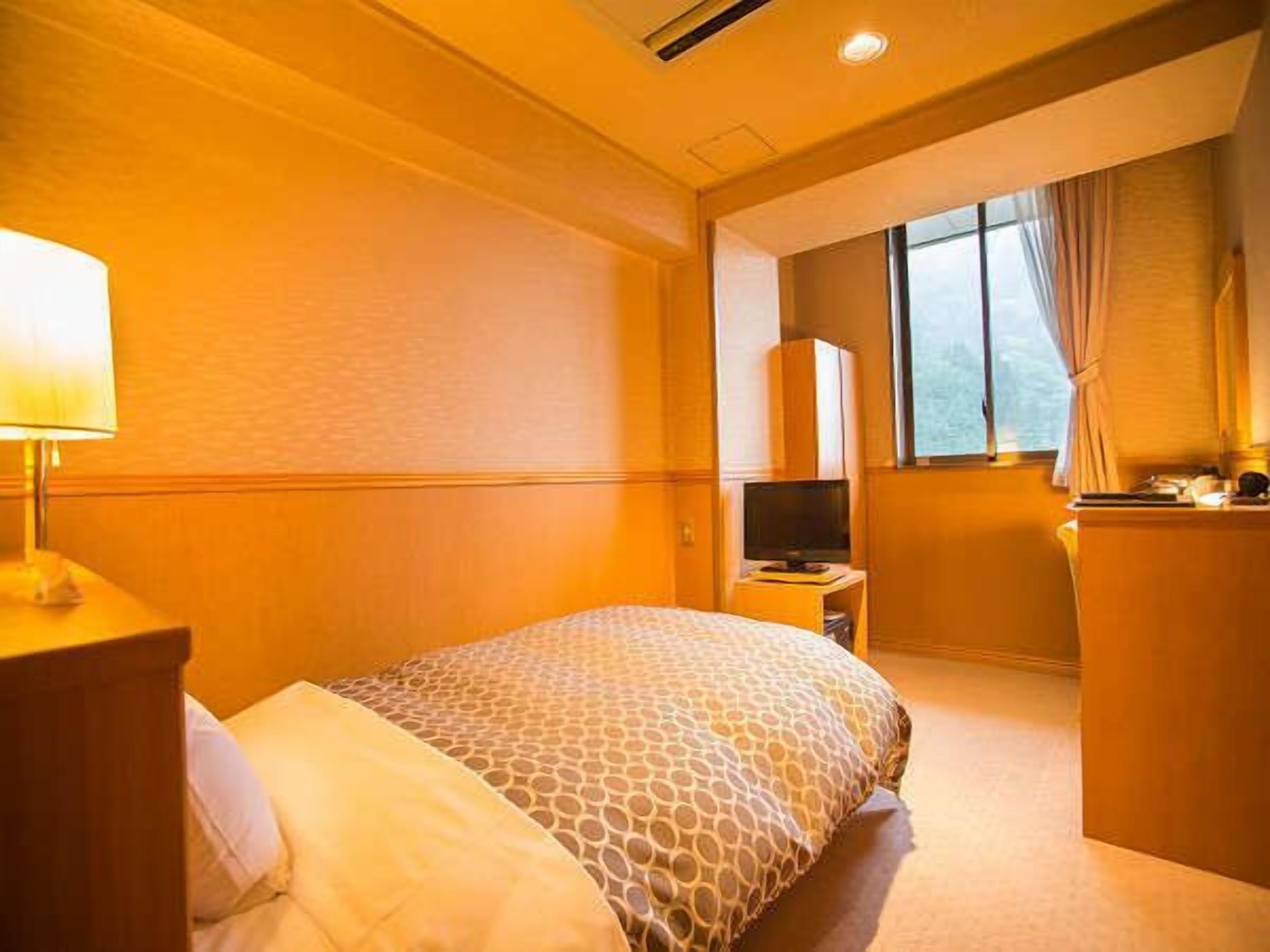 A hotel room with a bed, a desk with a lamp, a television, and a window with curtains.