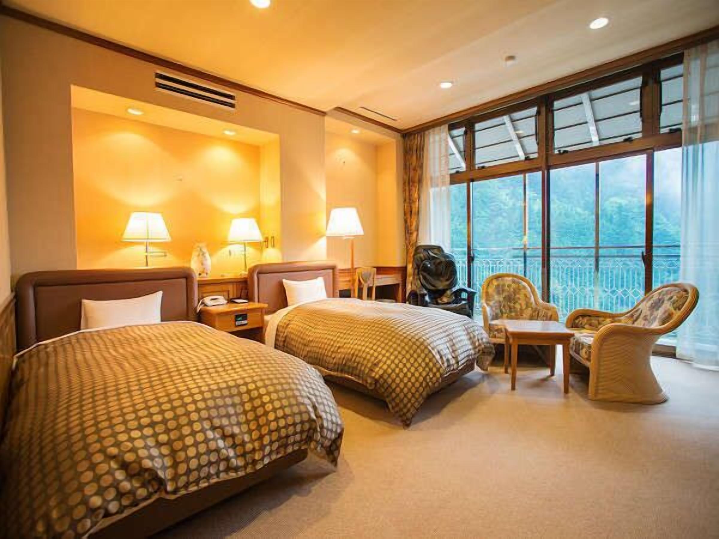 Camera d'albergo con due letti, un piccolo tavolo, due sedie, una lampada e vista sull'esterno.